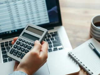 Hand with calculator beside laptop showing spreadsheet, coffee mug, notebook on wooden desk.